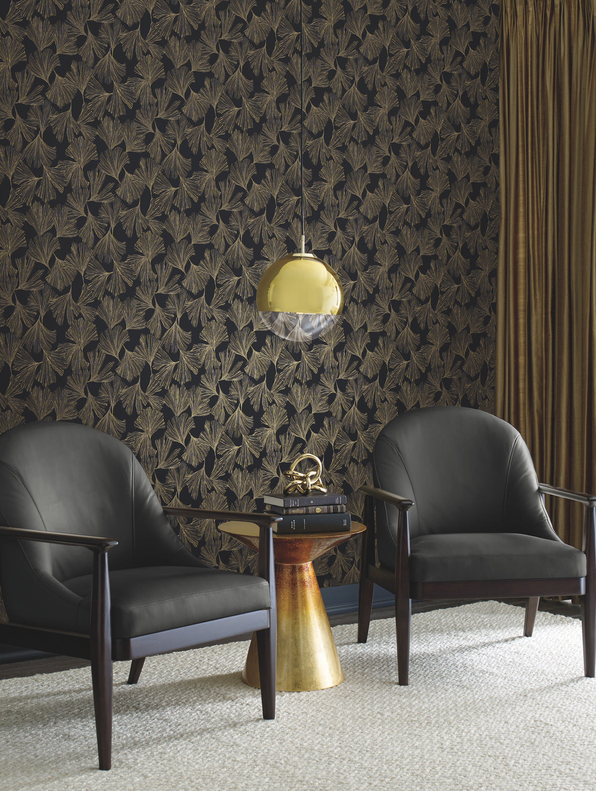 Two black armchairs face a small round gold side table with books and decor, set against a dark wall adorned with York Wallcoverings’ Gingko Toss Wallpaper. A gold pendant light hangs above, complemented by gold curtains on the right. A light rug covers the floor.
