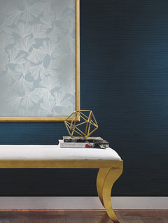A gold-bench with a white cushion holds books and a gold geometric sculpture. Behind it, the wall is covered with York Wallcoverings’ Gingko Toss Wallpaper featuring light leaf patterns framed as artwork, while the floor is medium-toned wood.