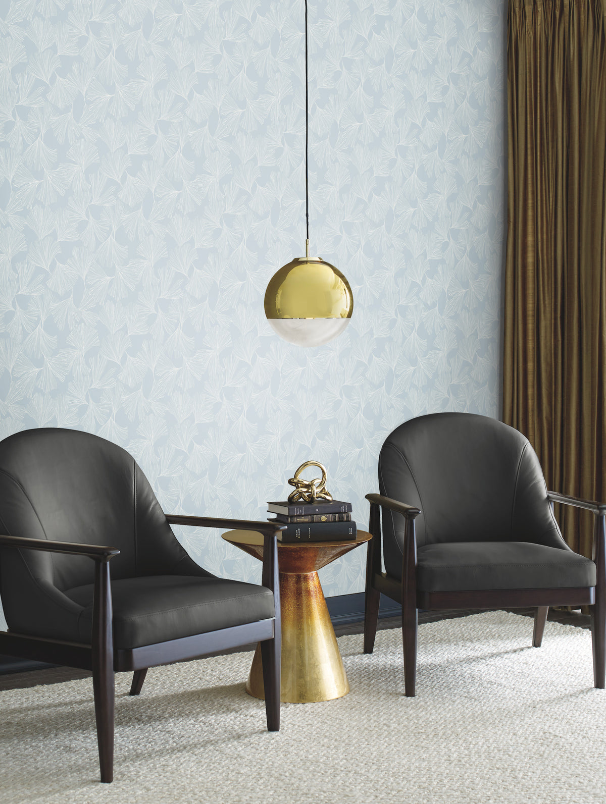 Two black armchairs with wooden arms flank a gold side table topped with books and a brass sculpture, beneath a gold hanging lamp. The background features York Wallcoverings’ Gingko Toss Wallpaper and brown curtains.