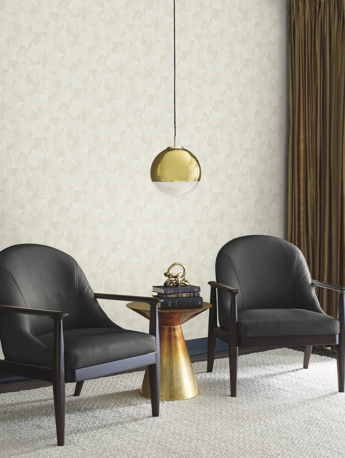 Two black armchairs sit beside a small gold side table with books and a sculpture. A gold pendant lamp hangs above. Gingko Toss Wallpaper by York Wallcoverings in beige covers the background, complemented by brown curtains. A textured rug covers the floor.