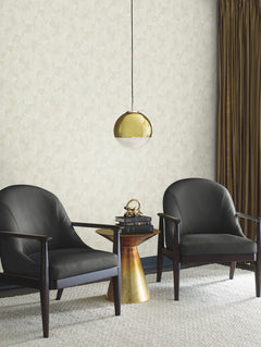 Two black armchairs sit beside a small gold side table with books and a sculpture. A gold pendant lamp hangs above. Gingko Toss Wallpaper by York Wallcoverings in beige covers the background, complemented by brown curtains. A textured rug covers the floor.