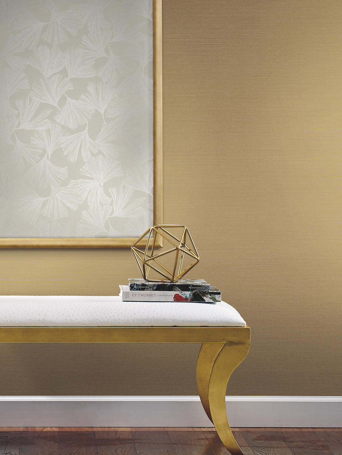 A white cushioned bench with gold legs sits against a wall adorned with Gingko Toss Wallpaper by York Wallcoverings. On the bench are two books and a gold geometric sculpture. Above the bench hangs framed, leaf-patterned artwork.