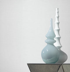 Two modern vases, one glossy light blue with a curvy shape and one matte white with a segmented design, sit on a geometric table against a plain, textured white Gossamer Woven Wallpaper by York Wallcoverings.