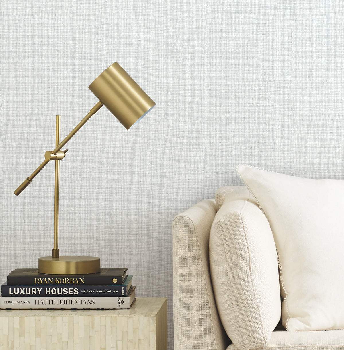 A gold desk lamp sits on a small table stacked with books next to a beige sofa with a cream pillow, set against a light textured wall covered in Gossamer Woven Wallpaper by York Wallcoverings.