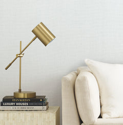 A gold desk lamp sits on a small table stacked with books next to a beige sofa with a cream pillow, set against a light textured wall covered in Gossamer Woven Wallpaper by York Wallcoverings.