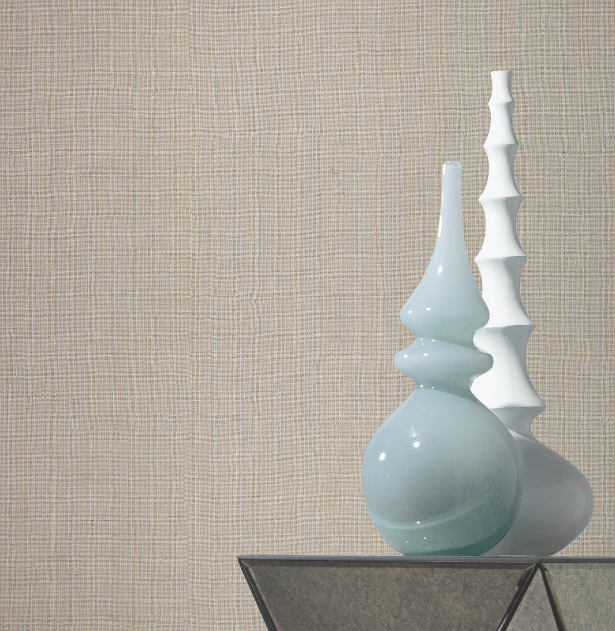 Two modern vases, one glossy light blue with curved shapes and one matte white with stacked segments, stand on a glass surface against a wall adorned with Gossamer Woven Wallpaper by York Wallcoverings.