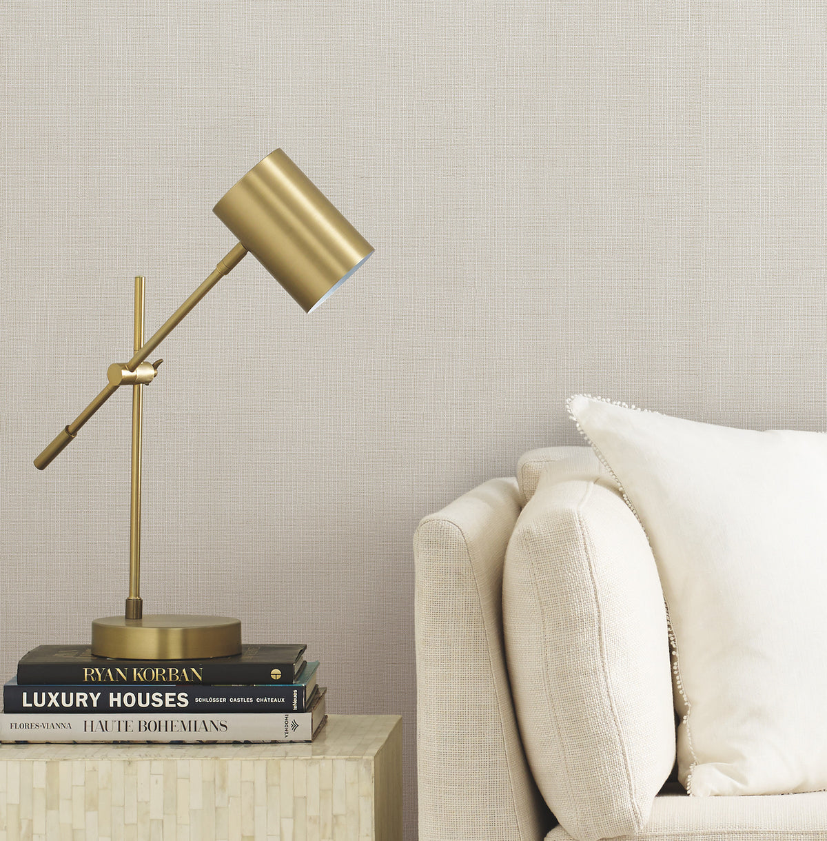 A brass desk lamp sits on a stack of books beside a beige sofa with a white pillow, against a wall adorned with York Wallcoverings’ Gossamer Woven Wallpaper.