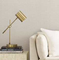 A brass desk lamp sits on a stack of books beside a beige sofa with a white pillow, against a wall adorned with York Wallcoverings’ Gossamer Woven Wallpaper.