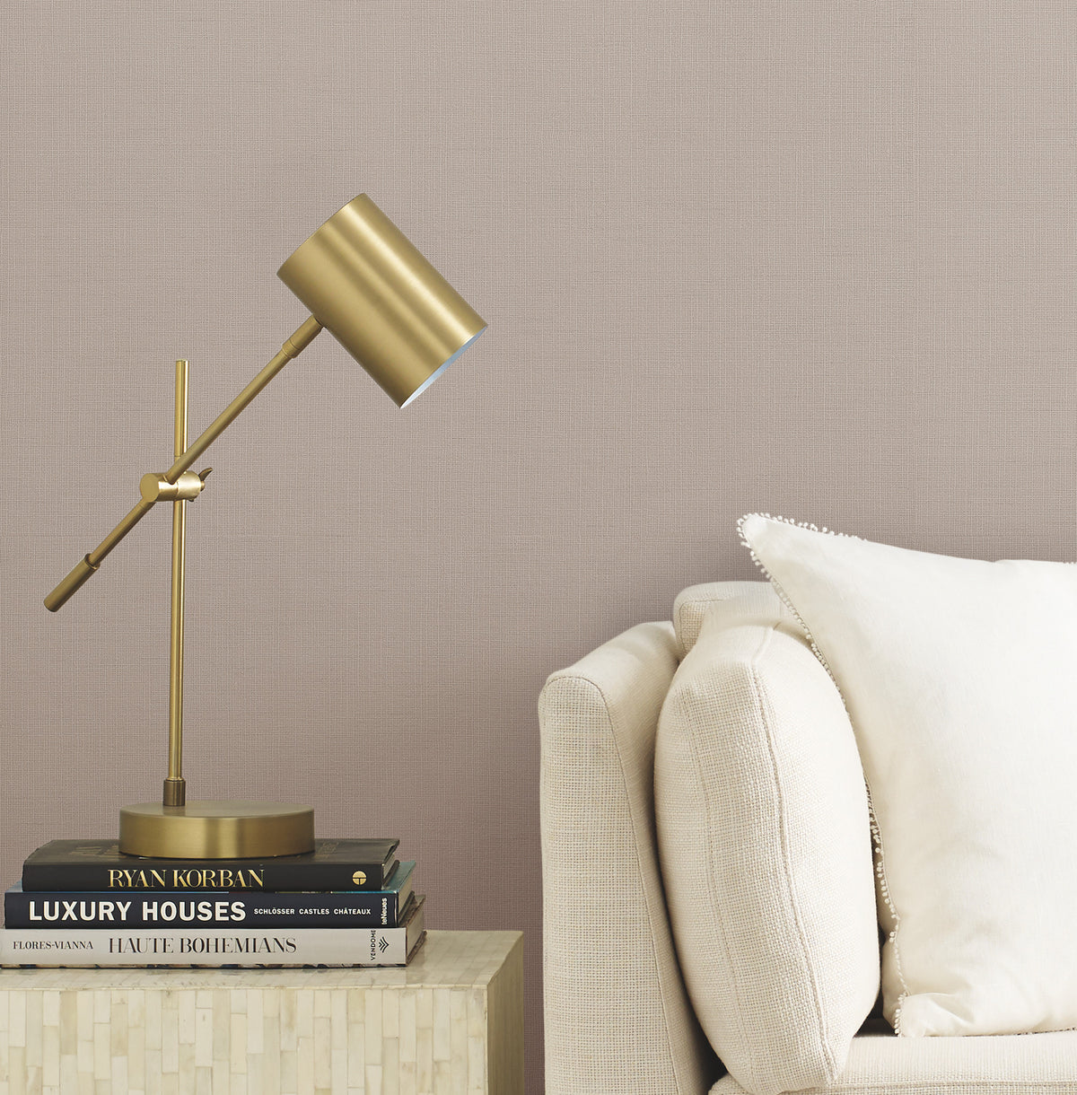 A gold desk lamp and stacked books sit on a light side table next to a cream-colored sofa with a white pillow, set against a wall adorned with Gossamer Woven Wallpaper by York Wallcoverings.