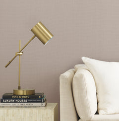 A modern brass desk lamp sits on a small table next to a beige sofa with a white pillow. Several stacked books are under the lamp. The background features Gossamer Woven Wallpaper by York Wallcoverings.