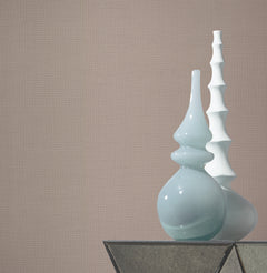 Two decorative vases sit on a glass surface—a light blue one with smooth, curved shapes and a white one with a tiered, spiked design—set against a plain beige background featuring Gossamer Woven Wallpaper by York Wallcoverings.