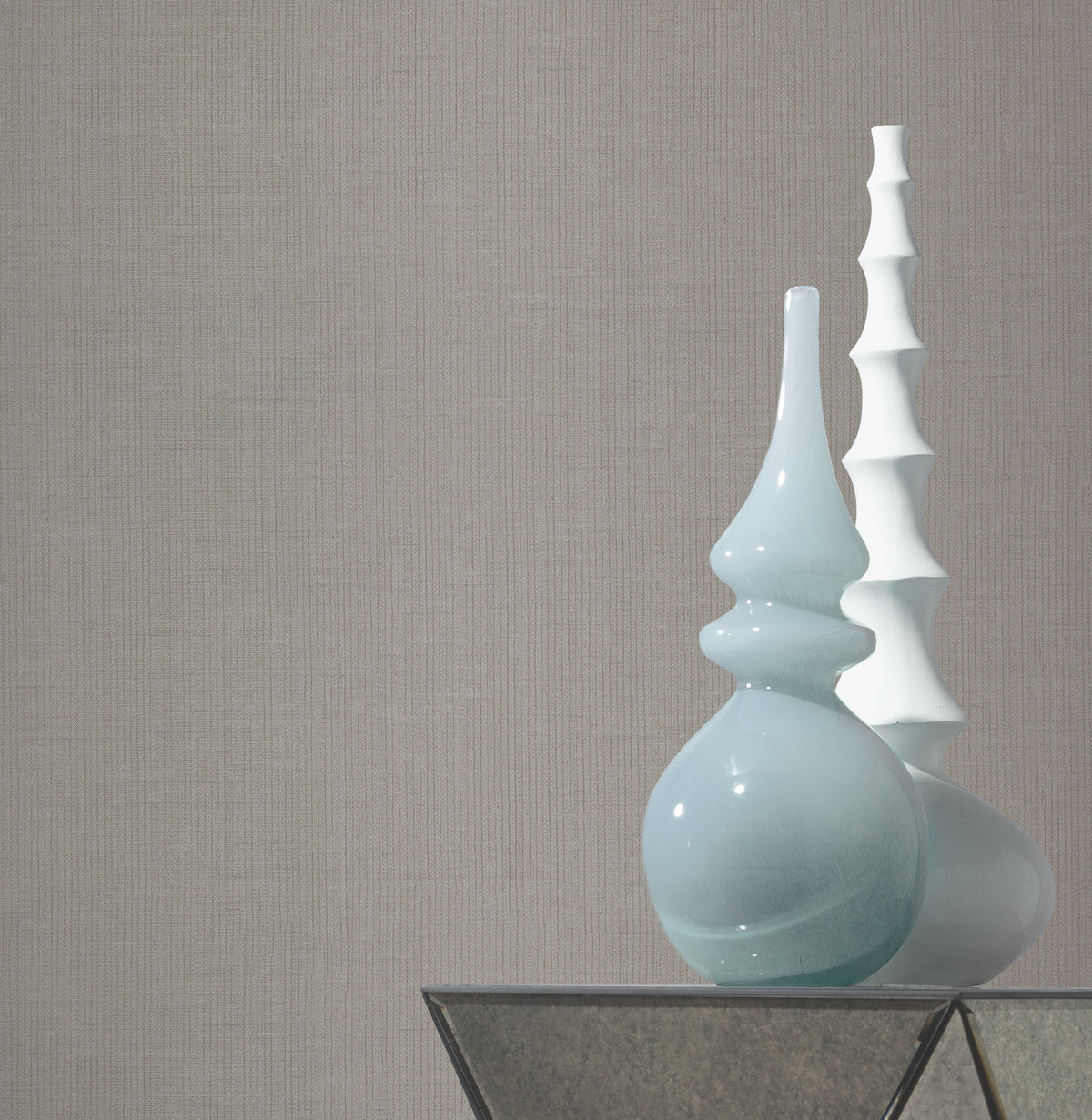 Two decorative vases, one light blue and curvy, the other white and spiky, sit on a glass-topped table against a wall adorned with York Wallcoverings’ Gossamer Woven Wallpaper.