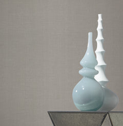 Two decorative vases, one light blue and curvy, the other white and spiky, sit on a glass-topped table against a wall adorned with York Wallcoverings’ Gossamer Woven Wallpaper.
