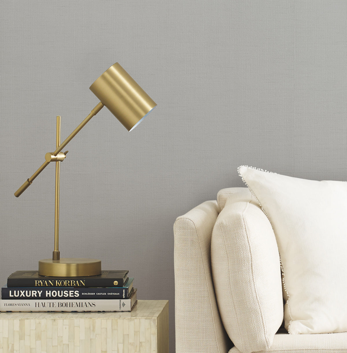 A gold desk lamp rests on a stack of books atop a small table beside a cream-colored sofa with a white pillow, set against a light gray Gossamer Woven Wallpaper by York Wallcoverings.