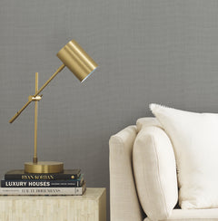 A modern gold desk lamp on a stack of design books sits next to a light beige sofa with a white pillow, set against a wall adorned with Gossamer Woven Wallpaper by York Wallcoverings.