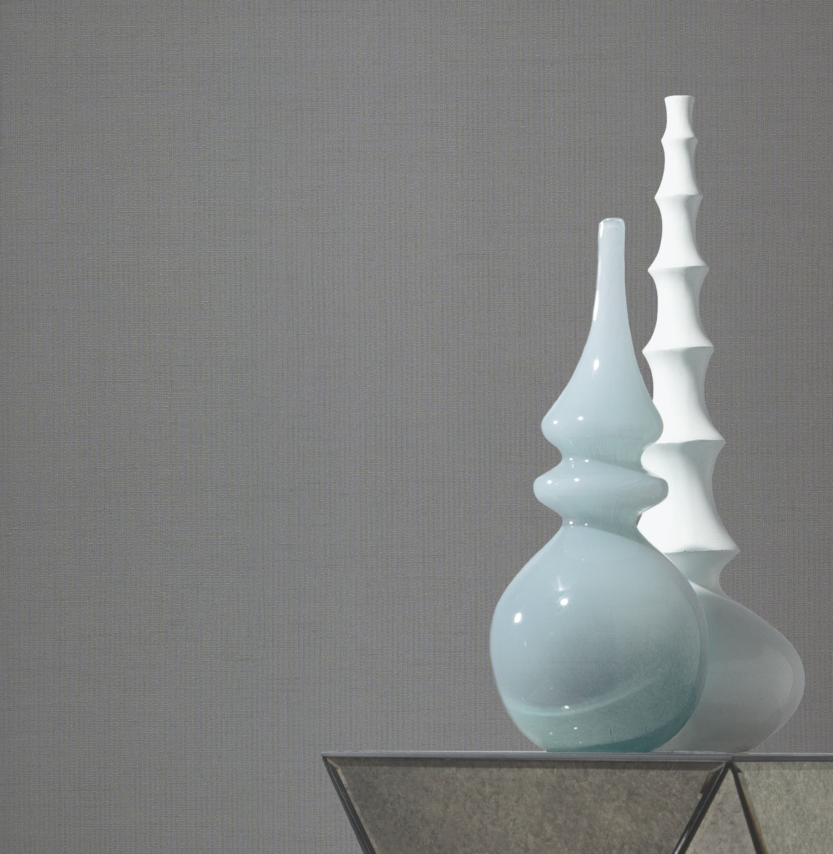 Two modern ceramic vases—one light blue and rounded, the other white with a wavy, tiered design—sit on a glass-topped table against a plain gray wall covered in Gossamer Woven Wallpaper by York Wallcoverings.