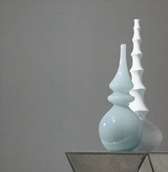 Two modern ceramic vases—one light blue and rounded, the other white with a wavy, tiered design—sit on a glass-topped table against a plain gray wall covered in Gossamer Woven Wallpaper by York Wallcoverings.