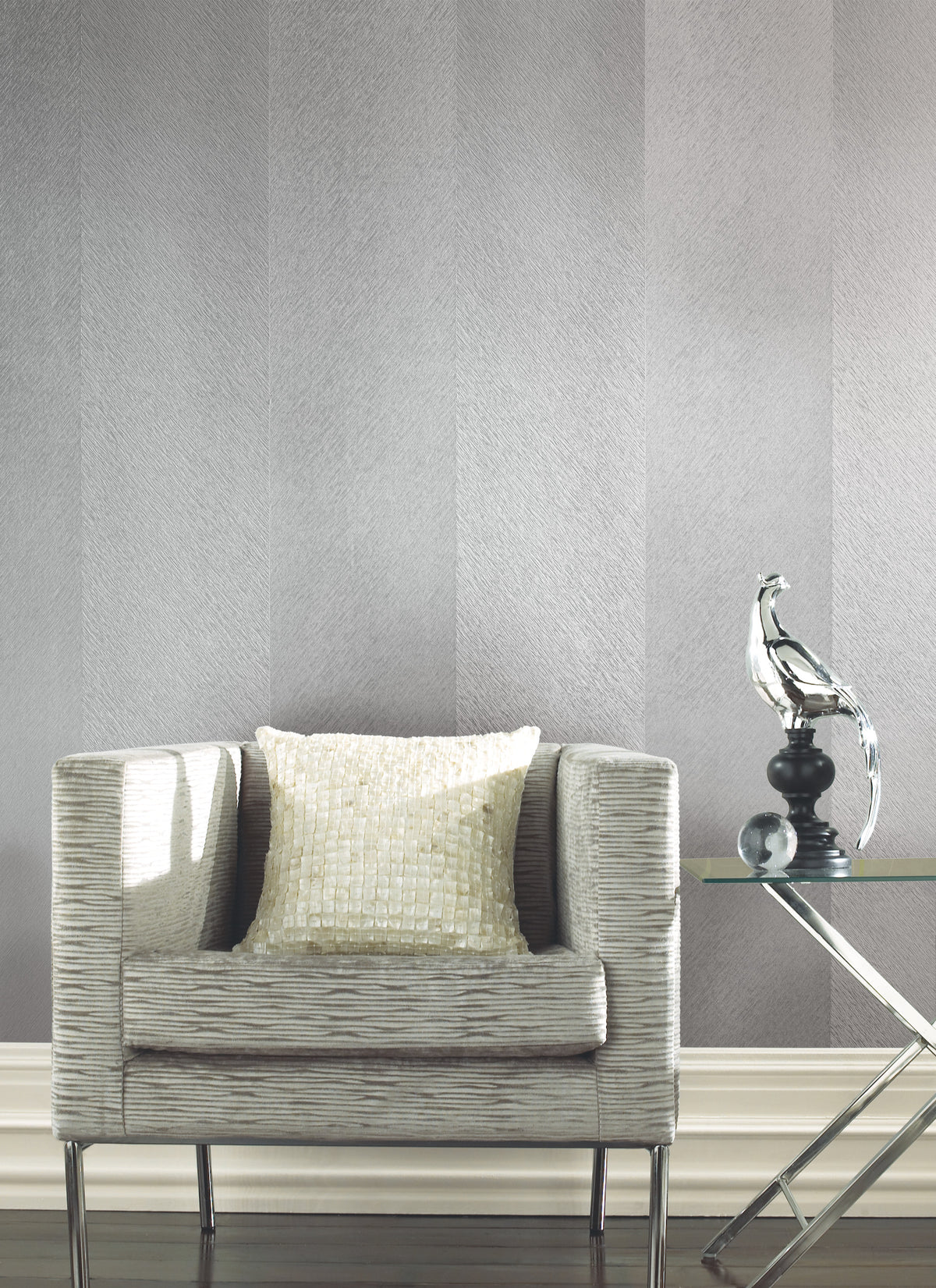 A modern armchair with a textured pillow sits beside a glass side table topped with a metallic bird sculpture, all set against York Wallcoverings’ Etched Chevron Wallpaper and dark wood flooring.