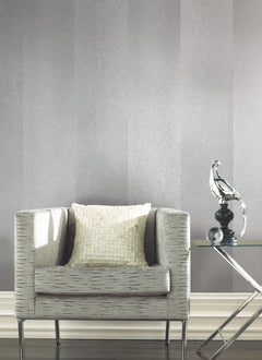 A modern gray armchair with a textured pillow sits next to a glass side table holding a silver decorative bird, set against York Wallcoverings’ Etched Chevron Wallpaper.