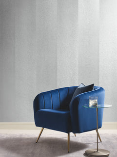 A modern blue velvet armchair with a cushion sits next to a round glass side table holding a glass of water, placed on a light gray rug against a wall adorned with Etched Chevron Wallpaper by York Wallcoverings.