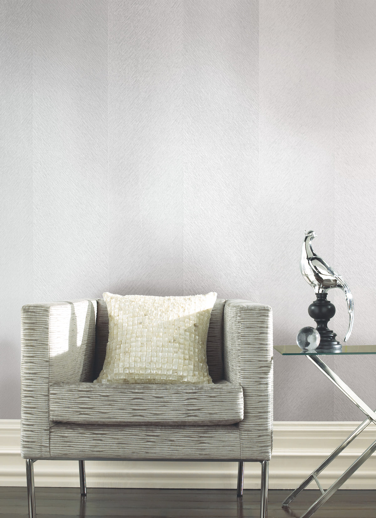 A modern beige armchair with a textured cushion sits beside a glass side table topped with a glass bird sculpture, set against York Wallcoverings’ Etched Chevron Wallpaper and a dark wood floor.