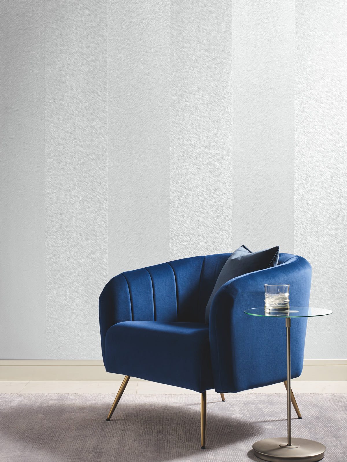 A modern blue armchair with a pillow sits beside a round glass side table holding a glass of water, on a light gray rug against a wall featuring the Etched Chevron Wallpaper by York Wallcoverings.