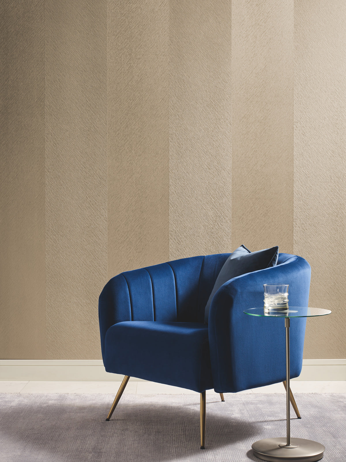 A blue velvet armchair with a cushion sits on a gray rug next to a round glass side table holding a glass of water, set against a wall covered in York Wallcoverings’ Etched Chevron Wallpaper.