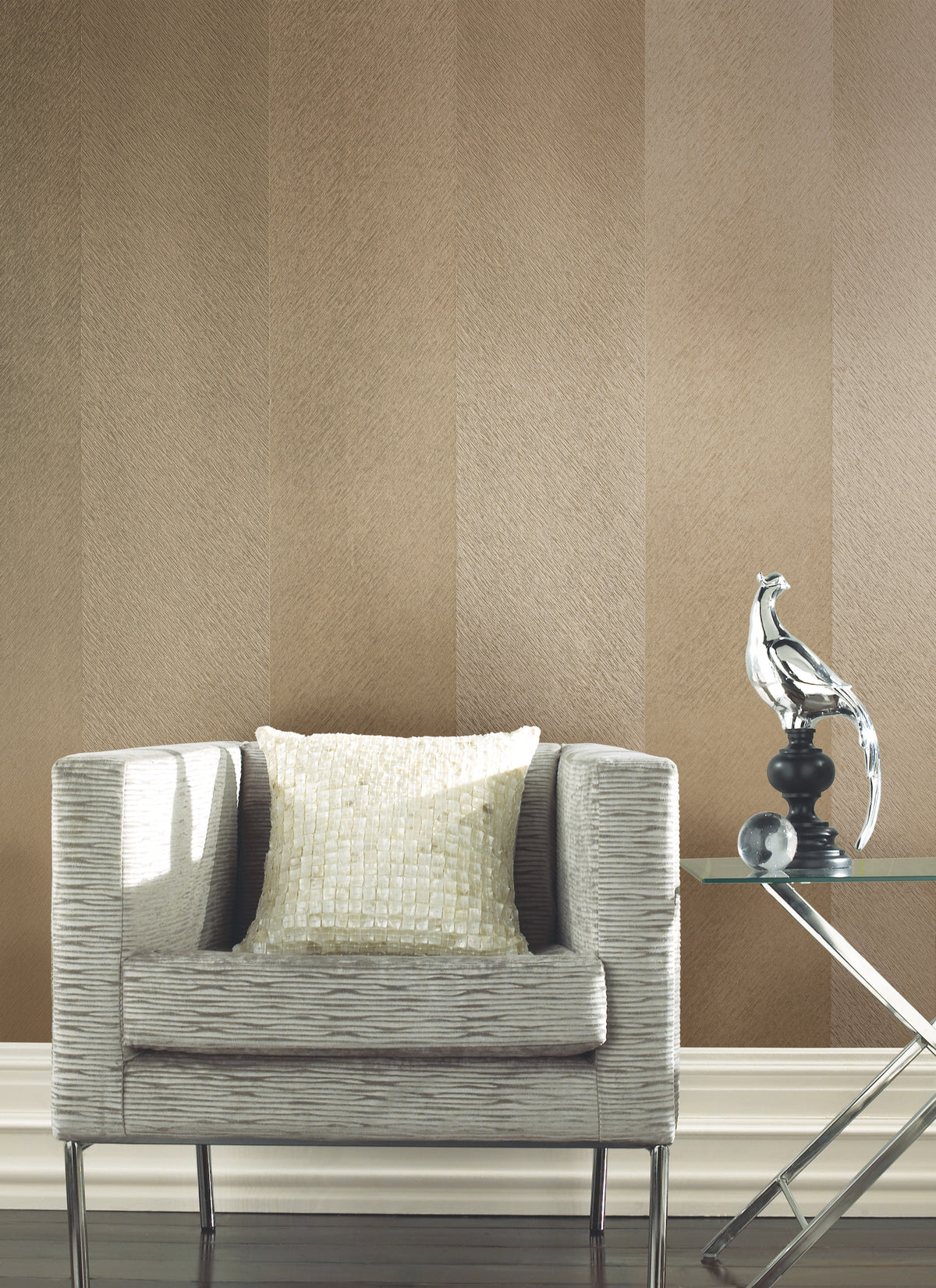 A modern armchair with a light textured pillow sits next to a glass side table displaying a silver abstract bird sculpture, all set against York Wallcoverings’ Etched Chevron Wallpaper.