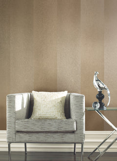 A modern armchair with a light textured pillow sits next to a glass side table displaying a silver abstract bird sculpture, all set against York Wallcoverings’ Etched Chevron Wallpaper.