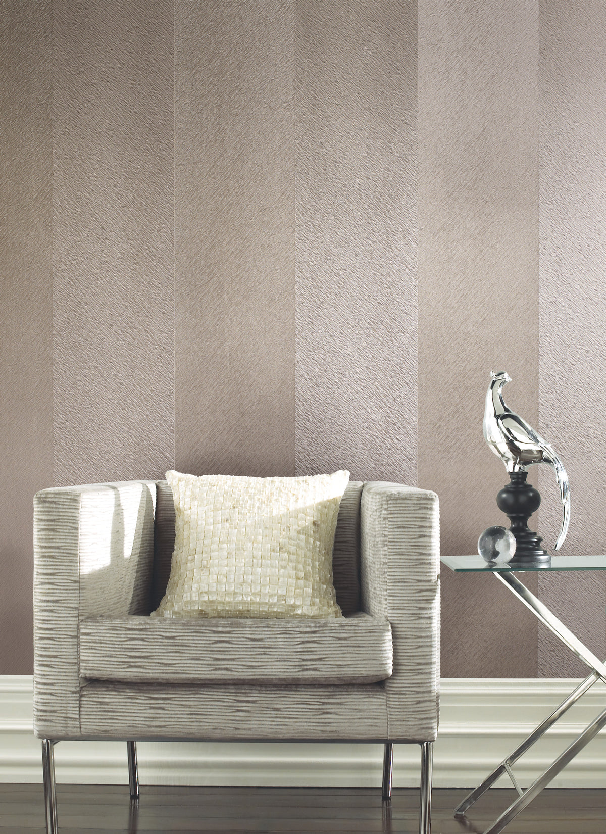 A textured beige armchair with a shiny cream pillow sits beside a glass side table holding a modern silver bird sculpture, set against walls covered in York Wallcoverings’ Etched Chevron Wallpaper.