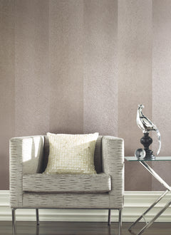 A textured beige armchair with a shiny cream pillow sits beside a glass side table holding a modern silver bird sculpture, set against walls covered in York Wallcoverings’ Etched Chevron Wallpaper.