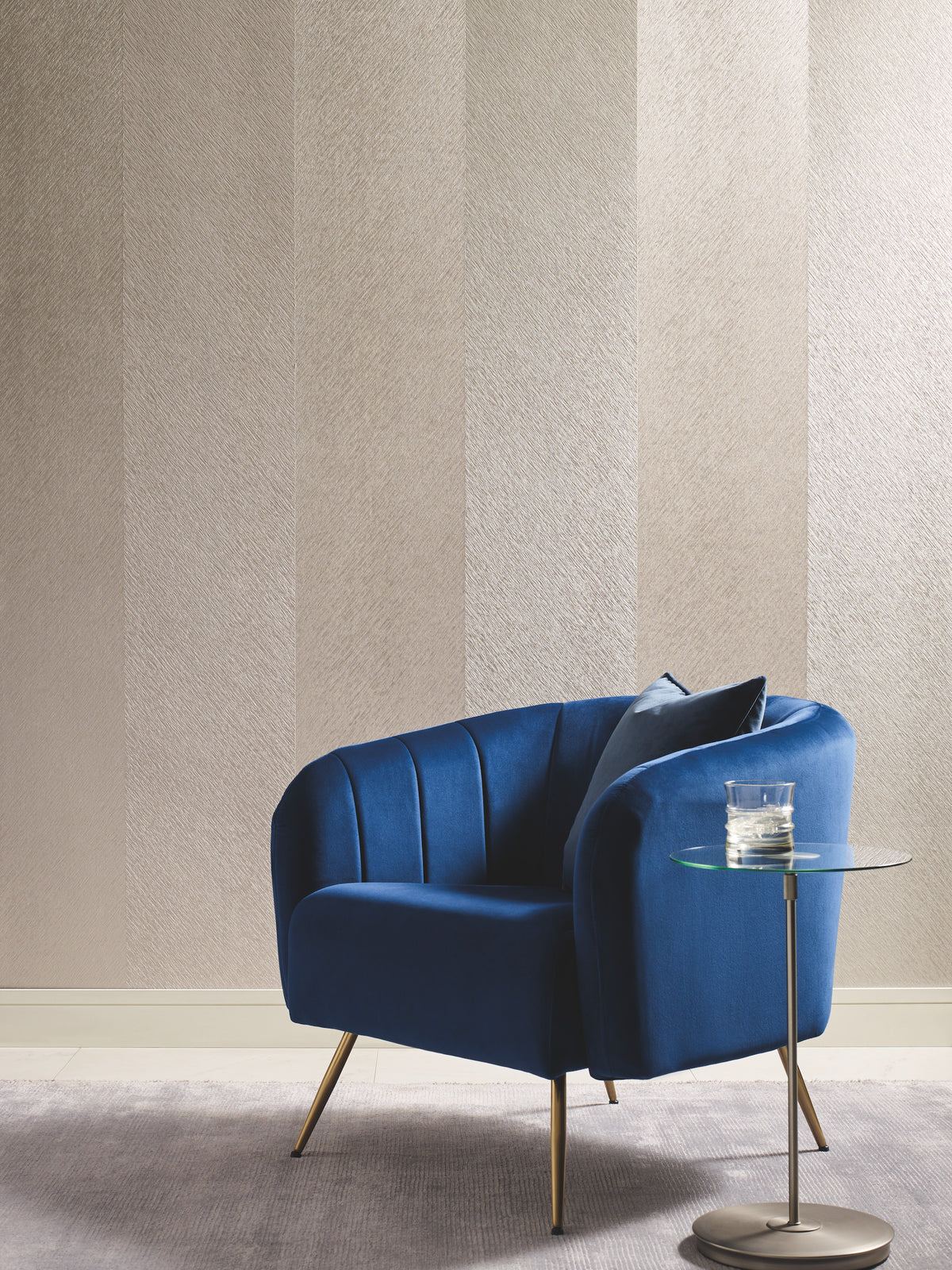 A modern blue armchair with gold legs sits on a gray rug, next to a round glass side table holding a glass of water, set against York Wallcoverings’ Etched Chevron Wallpaper.