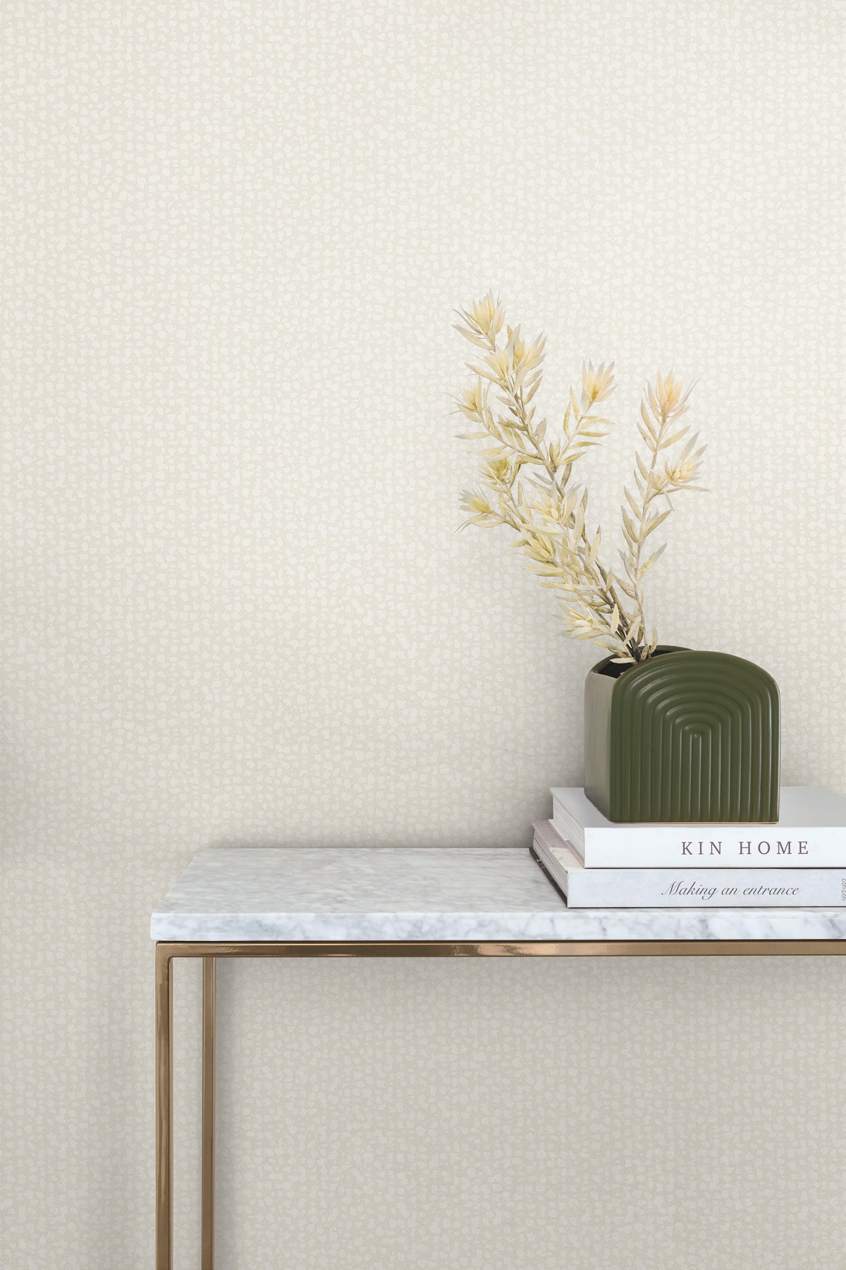 A console table with a marble top and gold legs stands against a wall covered in light, speckled Galaxies Wallpaper by York Wallcoverings, with a small vase of dried plants and two books on the surface.