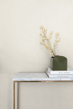 A console table with a marble top and gold legs stands against a wall covered in light, speckled Galaxies Wallpaper by York Wallcoverings, with a small vase of dried plants and two books on the surface.