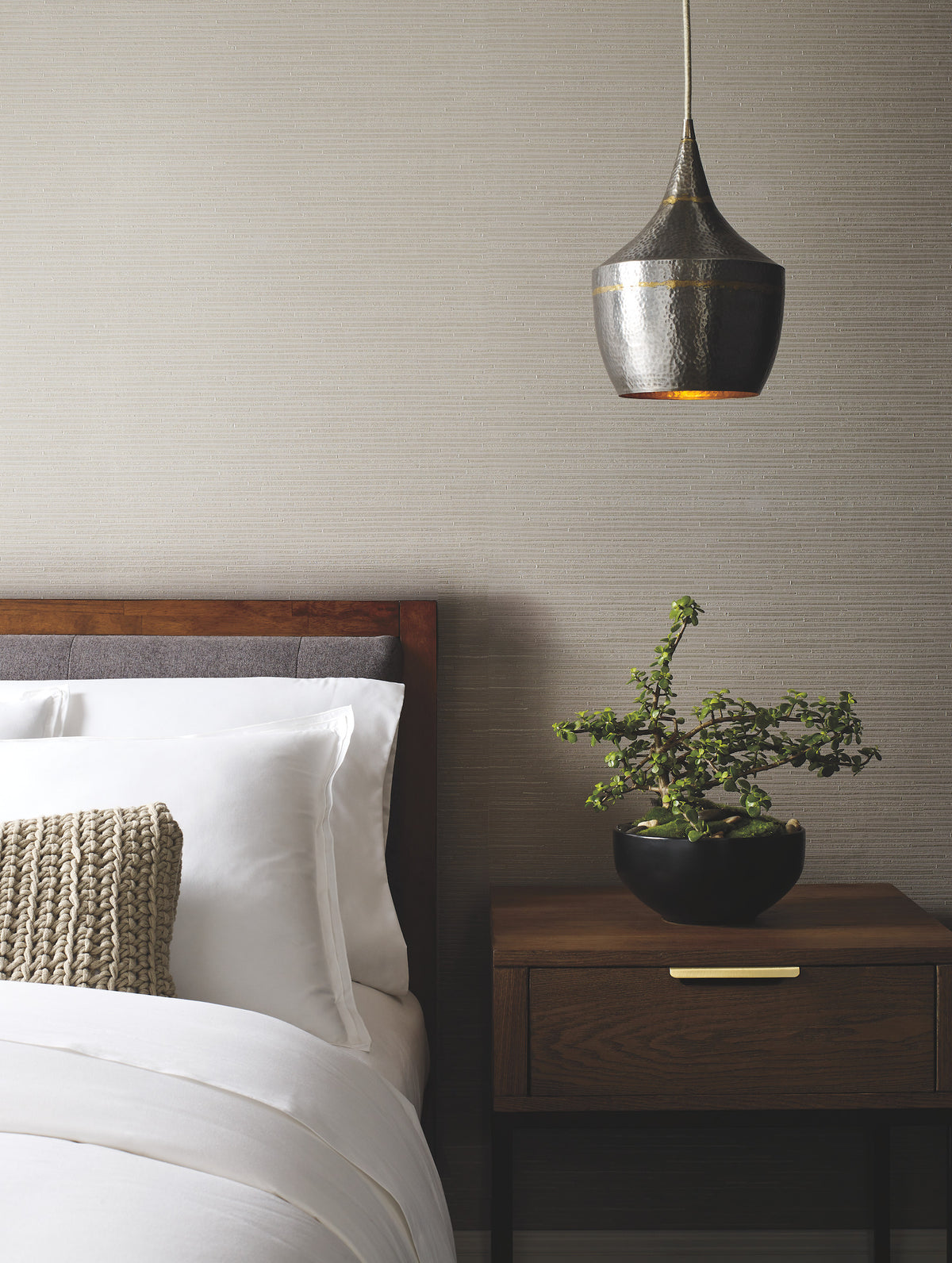 A neatly made bed with white linens and a textured pillow sits beside a wooden nightstand holding a potted plant, beneath a metallic hanging pendant light, against walls adorned with York Wallcoverings’ Ribbon Bamboo Wallpaper.