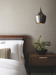 A neatly made bed with white linens and a textured pillow sits beside a wooden nightstand holding a potted plant, beneath a metallic hanging pendant light, against walls adorned with York Wallcoverings’ Ribbon Bamboo Wallpaper.