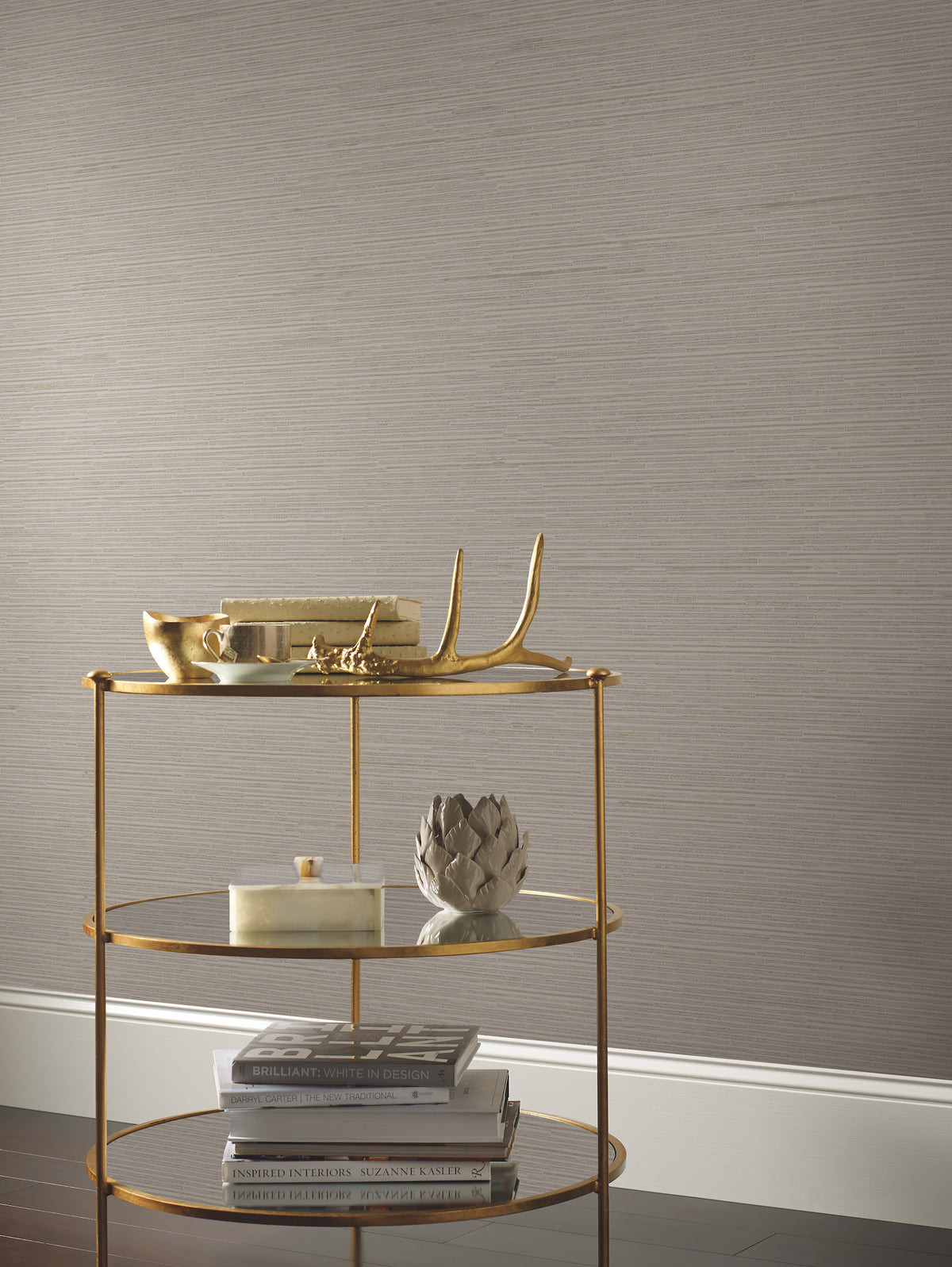 A gold three-tiered cart holds decorative items, books, and a sculpted artichoke against taupe Ribbon Bamboo Wallpaper by York Wallcoverings and a white baseboard.