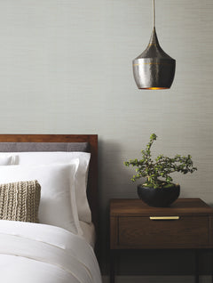 A modern bedroom features walls adorned with Ribbon Bamboo Wallpaper by York Wallcoverings, a wooden bed with white bedding, a knitted pillow, a wooden nightstand with a potted plant, and a dark metallic hanging pendant light above.