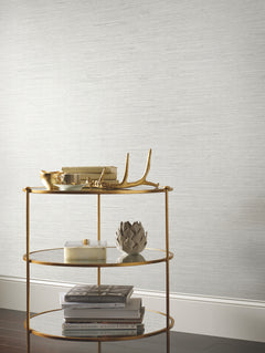 A gold bar cart holds decorative objects—including a candle, books, a gold antler-shaped piece, and a metallic artichoke—set against York Wallcoverings’ Ribbon Bamboo Wallpaper and a dark wood floor.