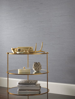 A gold three-tiered shelf holds decorative items and stacked books against York Wallcoverings’ Ribbon Bamboo Wallpaper, featuring a textured gray design with white trim at the base.