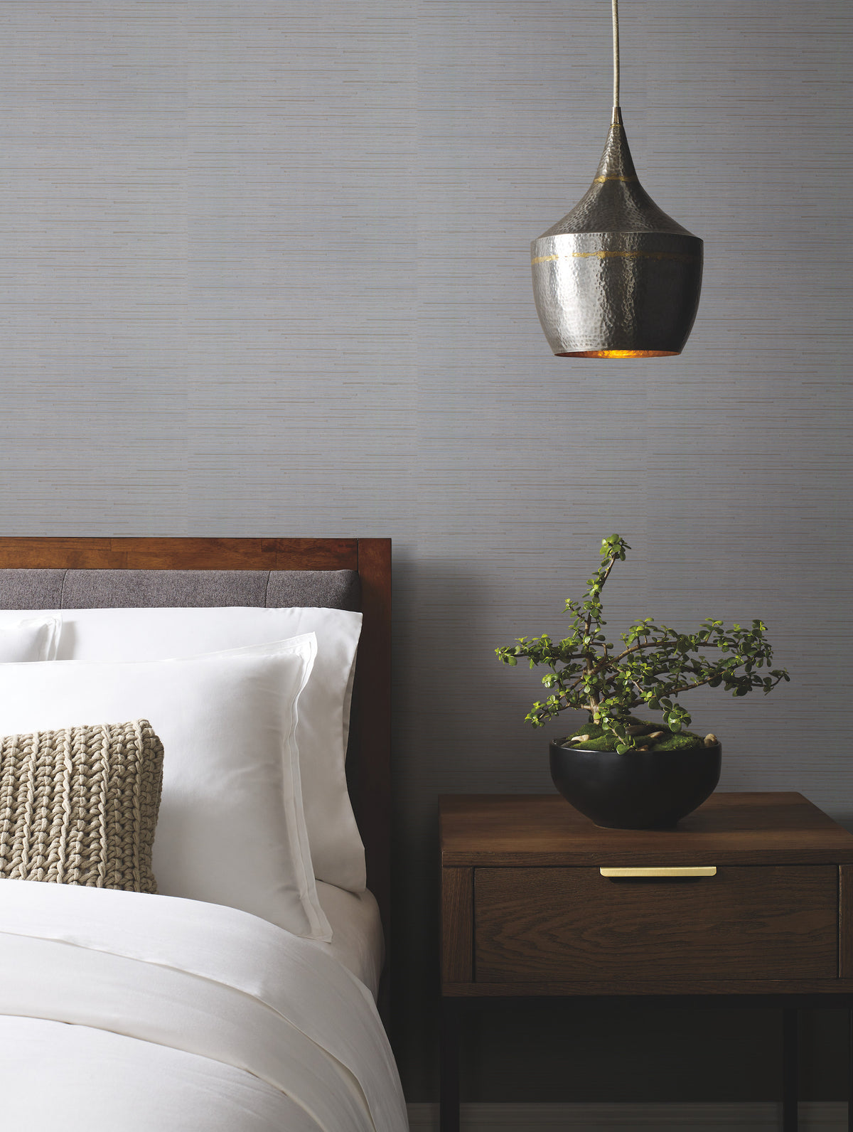 A modern bedroom featuring York Wallcoverings’ Ribbon Bamboo Wallpaper, a wooden bed with white bedding, a knitted cushion, a wooden nightstand adorned with a potted plant, and a metallic pendant light hanging above.