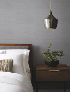 A modern bedroom featuring York Wallcoverings’ Ribbon Bamboo Wallpaper, a wooden bed with white bedding, a knitted cushion, a wooden nightstand adorned with a potted plant, and a metallic pendant light hanging above.
