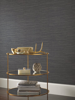 A gold bar cart adorned with decorative objects, books, and a small artichoke sculpture sits in front of a wall covered in York Wallcoverings’ Ribbon Bamboo Wallpaper, framed by white trim at the base.