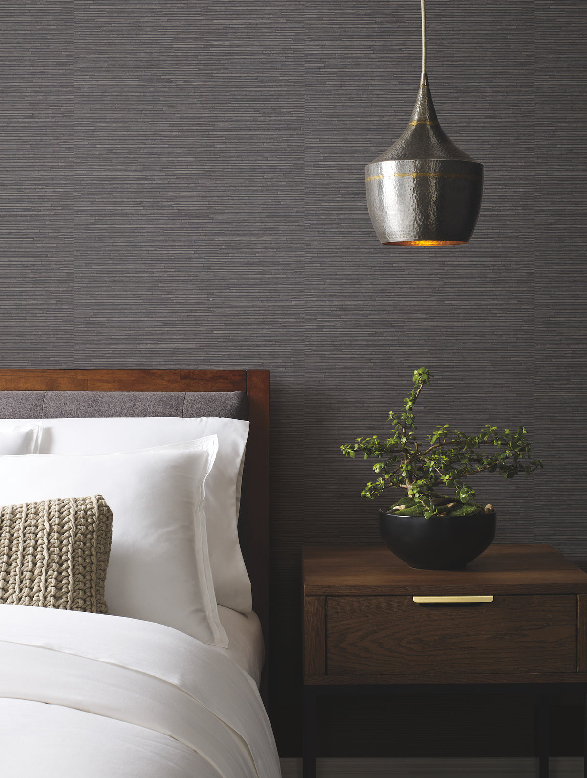 A modern bedroom features a wooden bed with white bedding, a knitted pillow, a wooden nightstand with a potted plant, and a metallic pendant light set against York Wallcoverings’ Ribbon Bamboo Wallpaper.