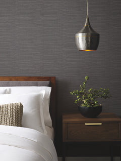 A modern bedroom features a wooden bed with white bedding, a knitted pillow, a wooden nightstand with a potted plant, and a metallic pendant light set against York Wallcoverings’ Ribbon Bamboo Wallpaper.