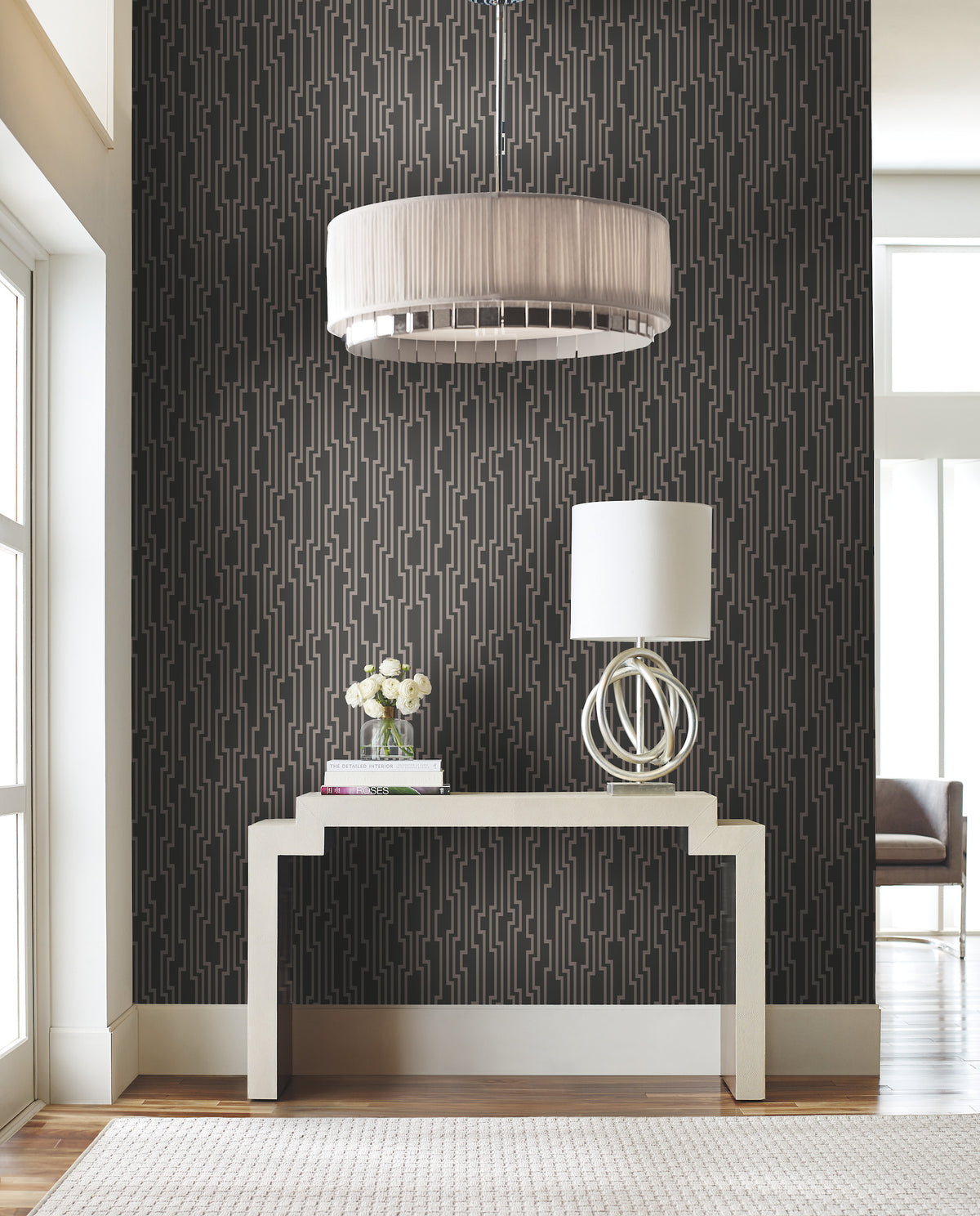 A console table with a lamp and vase of flowers against a wall covered in Velocity Wallpaper by York Wallcoverings, featuring a dark background with a geometric metallic line pattern.