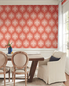 Dining room featuring a wooden table, beige upholstered chairs, a striped armchair, and York Wallcoverings’ Signet Medallion Damask Wallpaper in red. Sunlight streams through tall windows, illuminating a vase of flowers on the table.