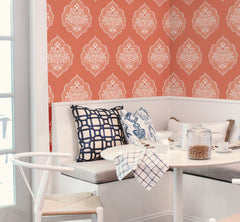 A cozy breakfast nook featuring a white bench, patterned pillows, a round white table set for breakfast with blue and white dishware, and York Wallcoverings’ Signet Medallion Damask Wallpaper in bold coral as the backdrop.
