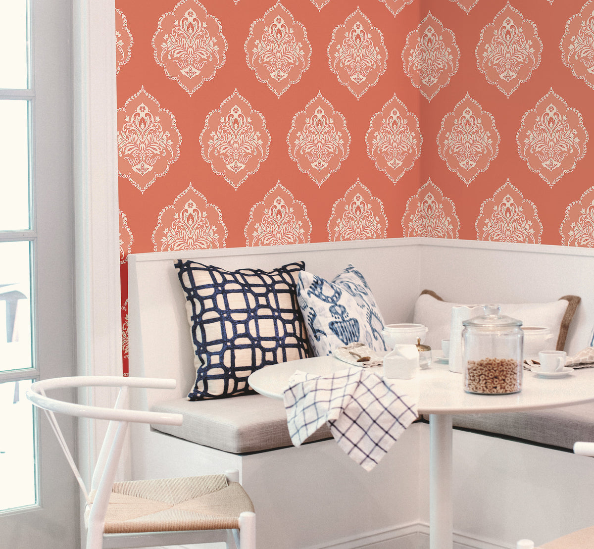 A cozy breakfast nook featuring a white bench, blue-patterned pillows, and a small table set for breakfast, with York Wallcoverings’ Signet Medallion Damask Wallpaper in a rich red pattern providing an elegant backdrop. Light streams in through a glass door on the left.