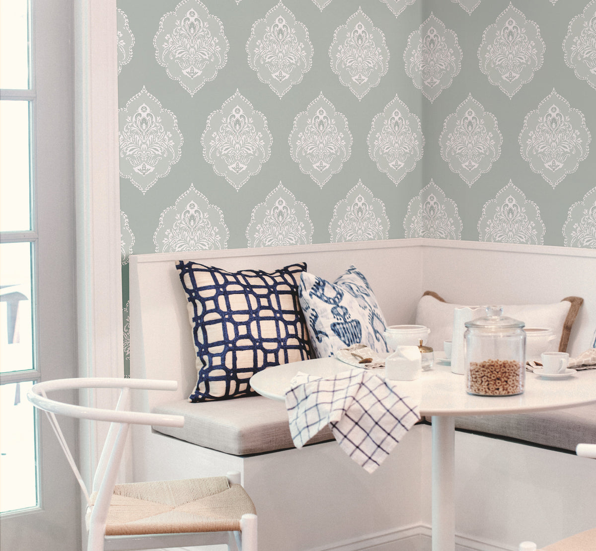 A cozy breakfast nook featuring a white corner bench, decorative blue and white pillows, a small round table, cereal jars, and a backdrop of York Wallcoverings’ Signet Medallion Damask Wallpaper.